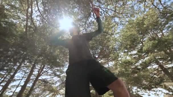 Молодой парень в шортах и кепке в солнечных очках идет балансировать на растянутой ленте в хвойном лесу во второй половине дня. Открытый спорт и досуг — стоковое видео
