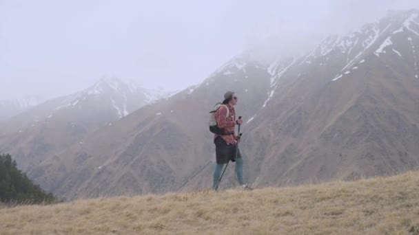 Un hombre alto joven en pantalones cortos camisa a cuadros en una gorra y mochila en gafas de sol va en una meseta alta contra el telón de fondo de las altas montañas en tiempo nublado. Seguimiento y descanso solitario en las montañas — Vídeos de Stock