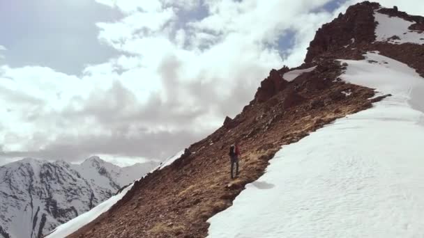 De klimmer klimt bergop met een rugzak en wandelstokken op de bergkam op de kruising van steen en sneeuw. Uitzicht vanuit de lucht hoog in de bergen van de noordelijke Kaukasus — Stockvideo