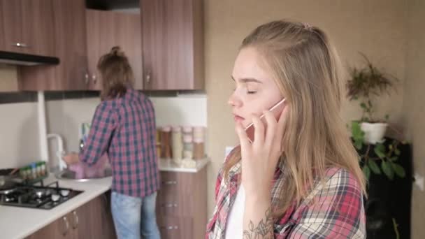 Disgruntled girl talking on the phone in the kitchen in the background of her boyfriend or husband. — Stock Video