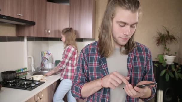 Serious elegante joven de pelo largo algo imprime en su teléfono inteligente en la cocina. Detrás de su espalda está su esposa o novia haciendo tareas alrededor de la casa — Vídeos de Stock