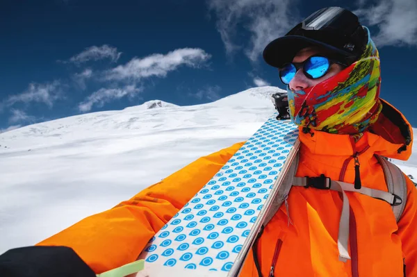 Retrato de um escalador de popa esquiador em óculos de sol e um boné com uma máscara de esqui em seu rosto. segura seus esquis em seu ombro e olha para o fundo do Monte Elbrus — Fotografia de Stock