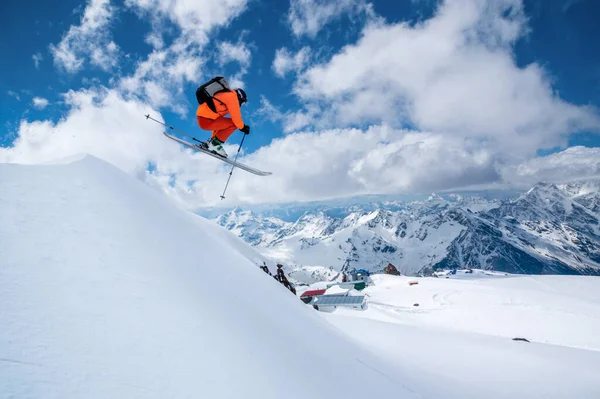 Ein männlicher Skifahrer in einem orangefarbenen Anzug fliegt in die Luft, nachdem er an einem sonnigen Tag zwischen schneebedeckten Bergen mit blauem Himmel und weißen Wolken von einer Schneedecke hoch in den kaukasischen Bergen gesprungen ist. — Stockfoto