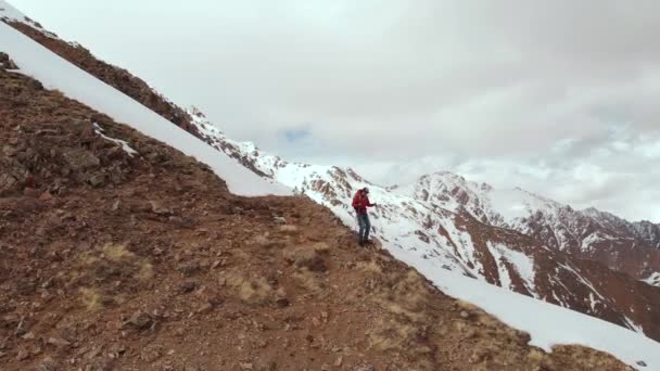 Luftbild junger Mann mit Sonnenbrille und Rucksack geht den Berg hinunter auf den Kamm an der Grenze zwischen Schnee und Steinen vor der Kulisse schneebedeckter Berge. — Stockvideo