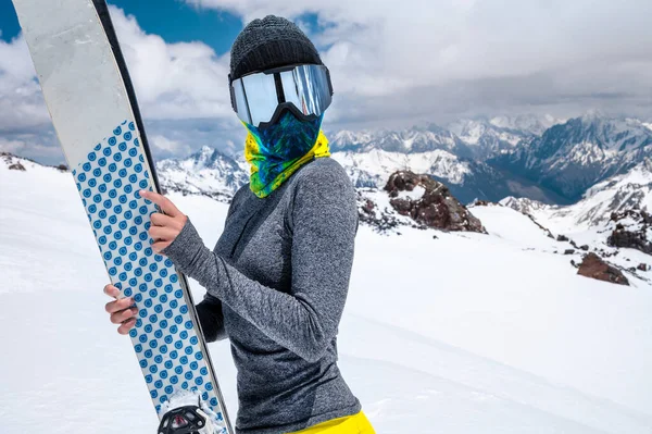 Portrait of a slender girl in a buff and balaclava in a ski mask and hat with a closed face next to skis on the background of snow-capped epic mountains.