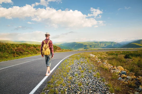 Un giovane hipster barbuto con un cappello a camicia e passeggiate a piedi nudi lungo una strada di campagna in montagna. Autostop e viaggi gratuiti — Foto Stock