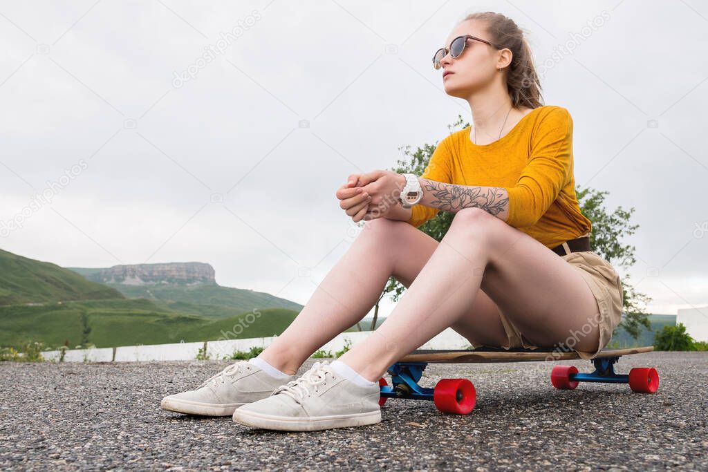 A young attractive girl in a yellow sweater shorts and sunglasses with a tattoo on her arm sits on a longboard behind a suburban asphalt pad