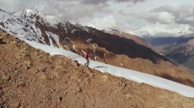 Sırt çantalı güneş gözlüklü genç adam kar ve taşlardan oluşan tepenin tepesinden kar örtülü dağlara doğru iniyor..
