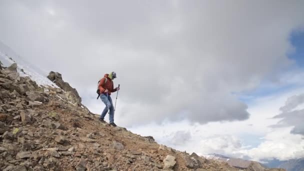 Een jonge man met een pet en een zonnebril met een rugzak en wandelstokken in zijn handen gaat de berg af tegen de achtergrond van hoge bergen — Stockvideo