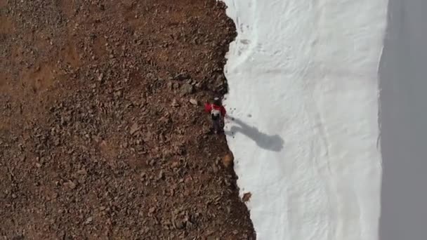 Vista de cima. O alpinista sobe colina acima com uma mochila e trekking sticks no cume na junção de pedra e neve. Vista aérea alta nas montanhas do norte do Cáucaso — Vídeo de Stock