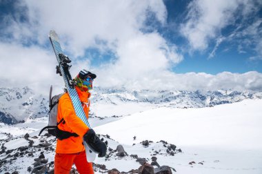 Güneş gözlüklü sert bir kayakçı portresi ve yüzünde kar maskesi olan bir şapka. Kayaklarını omzunda tutuyor ve Elbrus Dağı 'nın arka planına bakıyor.