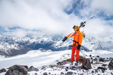 Güneş gözlüklü sert bir kayakçı portresi ve yüzünde kar maskesi olan bir şapka. Kayaklarını omzunda tutuyor ve Elbrus Dağı 'nın arka planına bakıyor.