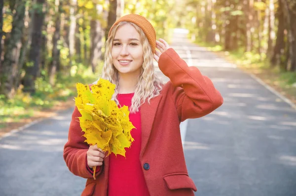 Jovem mulher atraente em um chapéu e casaco com um buquê de folhas amarelas caminha na floresta de outono em um dia ensolarado — Fotografia de Stock