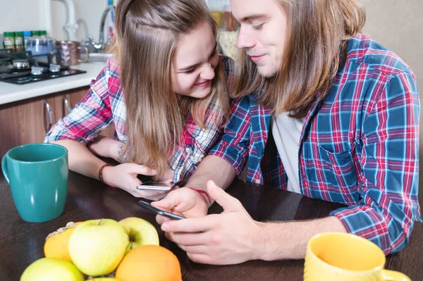 Young couple man and woman with long hair are sitting at the kitchen table in the kitchen and using their smartphones while buying food on the internet — Stock Photo, Image