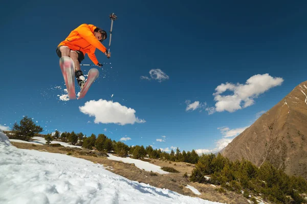 Um jovem homem elegante em óculos de sol e um boné executa um truque em saltar com um chute de neve contra o céu azul e o sol em um dia ensolarado . — Fotografia de Stock