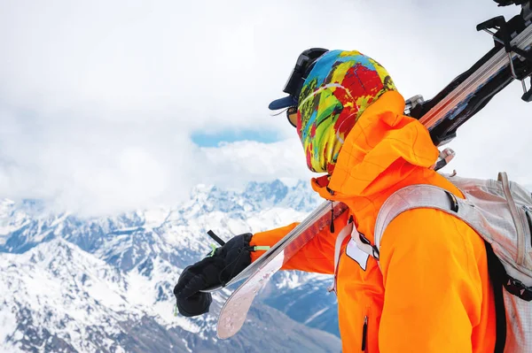 Ritratto di alpinista di poppa con occhiali da sole e berretto con maschera da sci sul viso. tiene gli sci sulla spalla e distoglie lo sguardo sullo sfondo del Monte Elbrus — Foto Stock