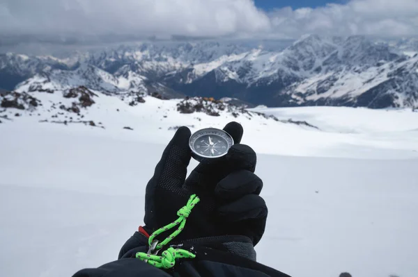 Una bussola metallica nella mano di un viaggiatore con un guanto nero sullo sfondo di montagne innevate — Foto Stock