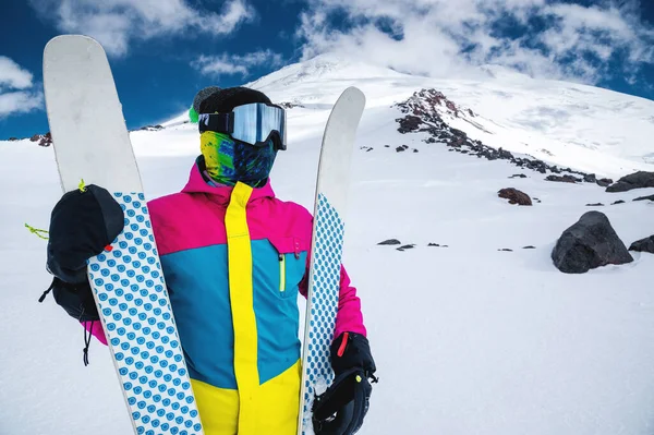 Porträt einer Skifahrerin in einer bunten Jacke mit Skimaske und geschlossenem Gesicht an einem sonnigen Tag vor dem Hintergrund schneebedeckter kaukasischer Berge und Wolken — Stockfoto