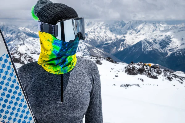 Retrato de uma mulher esquiadora sem casaco em uma máscara de esqui com o rosto fechado em um dia ensolarado contra o pano de fundo de montanhas e nuvens caucasianas cobertas de neve — Fotografia de Stock