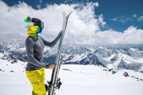 Porträt einer Skifahrerin ohne Jacke in Skimaske mit geschlossenem Gesicht an einem sonnigen Tag vor dem Hintergrund schneebedeckter kaukasischer Berge und Wolken — Stockfoto