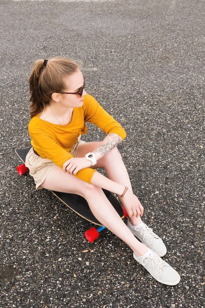 Uma jovem garota atraente em um suéter amarelo shorts e óculos de sol com uma tatuagem no braço senta-se em um longboard atrás de uma almofada de asfalto suburbana — Fotografia de Stock