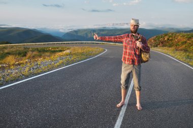 Tişört şapkalı ve çıplak ayaklı genç sakallı bir hippi dağlarda bir köy yolunda yürüyor. Otostop ve bedava seyahat