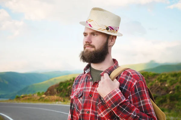 Retrato de um viajante sorridente feliz barbudo hipster com uma mochila em uma camisa xadrez e um chapéu ao lado de um carro desconhecido fica na estrada ao pôr do sol nas montanhas. Conceito de viagem feliz e confiante — Fotografia de Stock