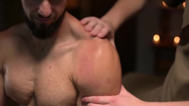 Un massaggiatore maschile fa un massaggio sportivo alle spalle a un atleta maschio muscoloso in una stanza con una luce scura a contrasto. Massaggio sportivo professionale — Video Stock