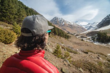 Uzun saçlı, kırmızı ceketli ve güneş gözlüklü gri şapkalı genç adam sırtı kameraya dönük duruyor ve uzaktaki karlı zirveleri olan dağ geçidine bakıyor.