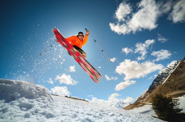 Güneş gözlüklü ve şapkalı genç şık bir adam güneşli bir günde mavi gökyüzüne ve güneşe karşı bir kar topuyla atlayarak bir numara yapıyor. Kışın veya ilkbaharda park kayağı konsepti