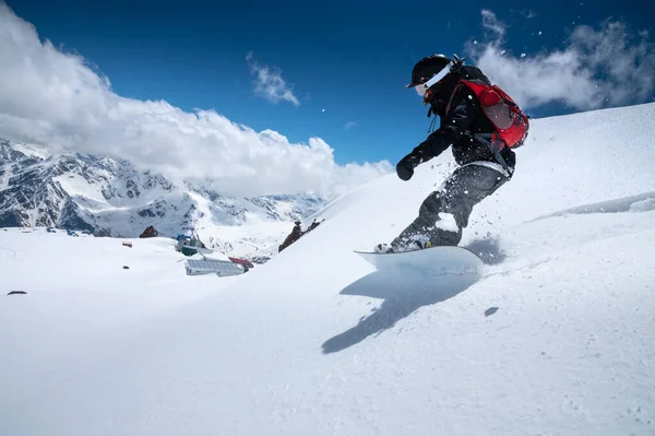 Сноубордистка с рюкзаком на снежном свежем склоне на фоне высоких гор и голубого неба. Зимние виды экстремального спорта. Сноуборд — стоковое фото