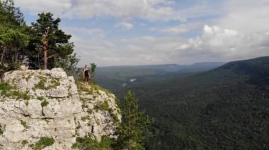 Siyah tişörtlü ve şortlu genç bir adam yüksek bir uçurumun kenarında duruyor ve etrafına bakıyor. Kaya ve ormanlı doğa rezervinin havadan görünüşü