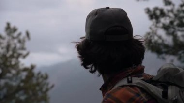 Dağların bulutlu havasında, akşamları ormandan çıkan, uzun saçlı, şapkalı genç bir adamın yakın görüntüsü. Seyahat ve seyahat.