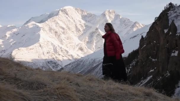 Atractiva joven con un vestido negro y chaqueta roja va cuesta arriba en una ladera cubierta de hierba contra las altas montañas nevadas del Cáucaso — Vídeo de stock