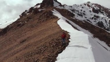 Sırt çantalı güneş gözlüklü genç adam kar ve taşlardan oluşan tepenin tepesinden kar örtülü dağlara doğru iniyor..