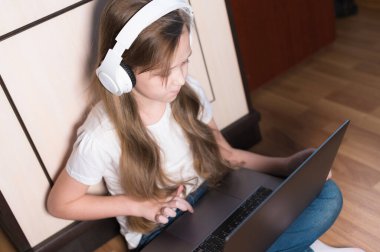 Kulaklıklı, ciddi bir suratı olan küçük bir kız kucağında dizüstü bilgisayarla yerde oturuyor ve ödevini yapıyor. Uzak öğrenme