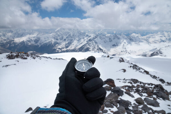 Magnetic compass in a mans hand in a glove against the background of the snow-capped mountains of the caucasus in the afternoon. Navigation concept