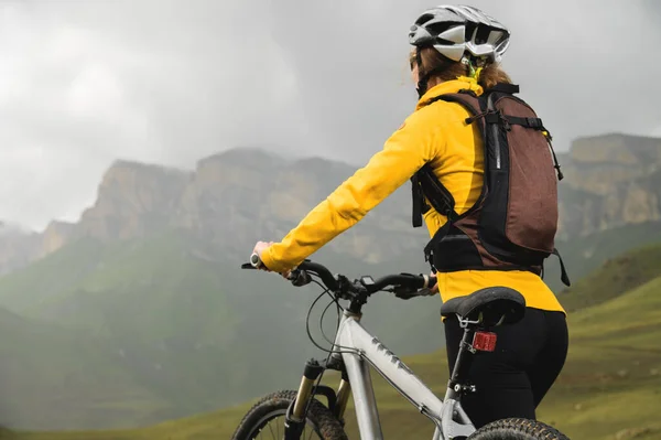 Una mujer joven en un casco y con una mochila se para con una bicicleta y mira a las montañas en un clima nublado. bicicleta de montaña mtb —  Fotos de Stock