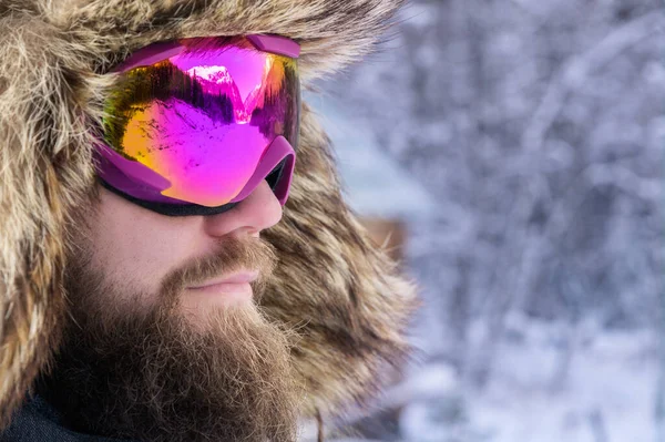 Κοντινό πλάνο πορτρέτο ενός ελκυστικού γενειοφόρου άνδρα με μάσκα του σκι και γούνινο καπέλο στο φόντο ενός χιονισμένου δάσους — Φωτογραφία Αρχείου