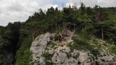 Siyah tişörtlü ve şortlu genç bir adam yüksek bir uçurumun kenarında duruyor ve etrafına bakıyor. Kaya ve ormanlı doğa rezervinin havadan görünüşü