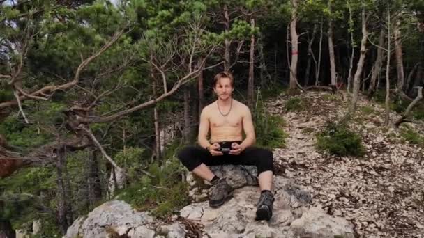 En attraktiv langhåret, atletisk mand sidder på kanten af en klippe holder en drone kontrolpanel. Drone flyvninger i naturreservater og drone rekreation – Stock-video