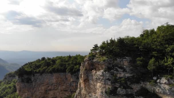 A young man in a black T-shirt and shorts stands on the edge of a cliff in front of a cliff against the backdrop of a forest mountain valley in summer. Aerial view — Stock Video