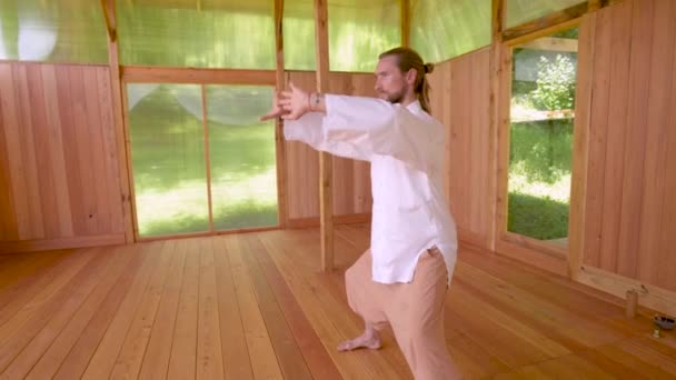 Un bărbat cu barbă caucaziană, cu părul blond lung, practică qigong și taichi într-o sală de practică din lemn din pădurea de vară. Practica artelor marțiale în teritoriile europene — Videoclip de stoc