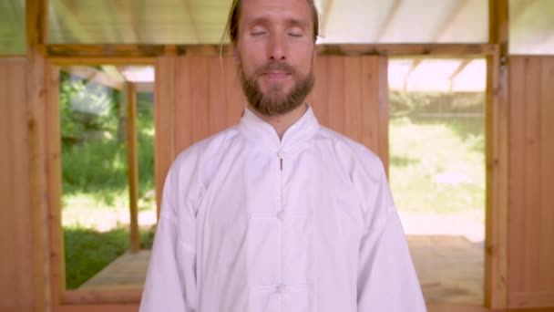 Gros plan Homme caucasien barbu portant des vêtements blancs pour pratiquer la concentration et la respiration avant de pratiquer le qigong tai chi — Video