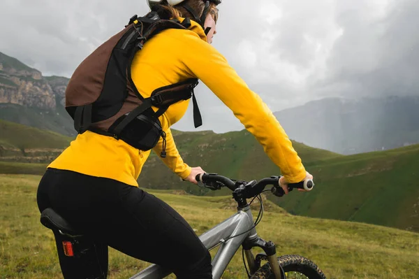 Una mujer joven en un casco y con una mochila se para con una bicicleta y mira a las montañas en un clima nublado. bicicleta de montaña mtb —  Fotos de Stock