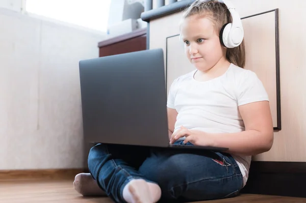 A small plump fat girl with a serious face in headphones sits on the floor in a room with a laptop in her arms and does her homework. Remote learning