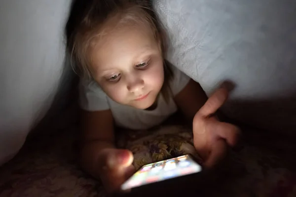 Geceleri yorganın altında akıllı telefonu olan küçük bir kız. Çocukluk aleti bağımlılığı ve uykusuzluk. — Stok fotoğraf