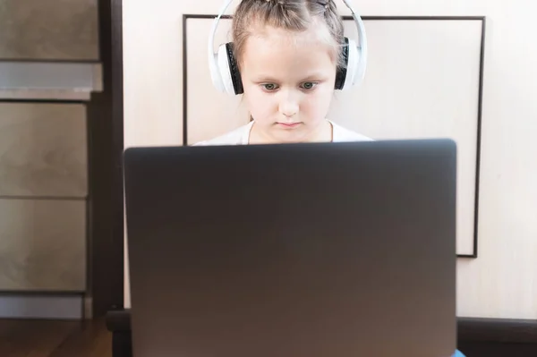 Een mollig dik meisje met een serieus gezicht in een koptelefoon zit op de grond in een kamer met een laptop in haar armen en doet haar huiswerk. Afstandsonderwijs — Stockfoto