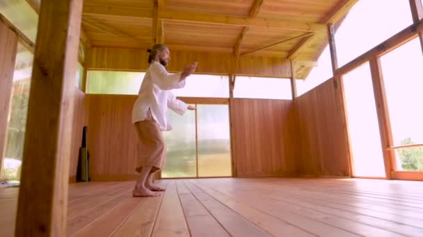 Een blanke baardman met lang blond haar oefent qigong en taichi uit in een houten oefenruimte in het zomerwoud. De beoefening van vechtsport op Europees grondgebied — Stockvideo