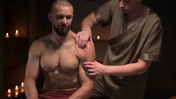 Manlig massör gör sport massage i lyx spa salong till manlig idrottsman med skägg på bakgrunden av brinnande ljus — Stockvideo